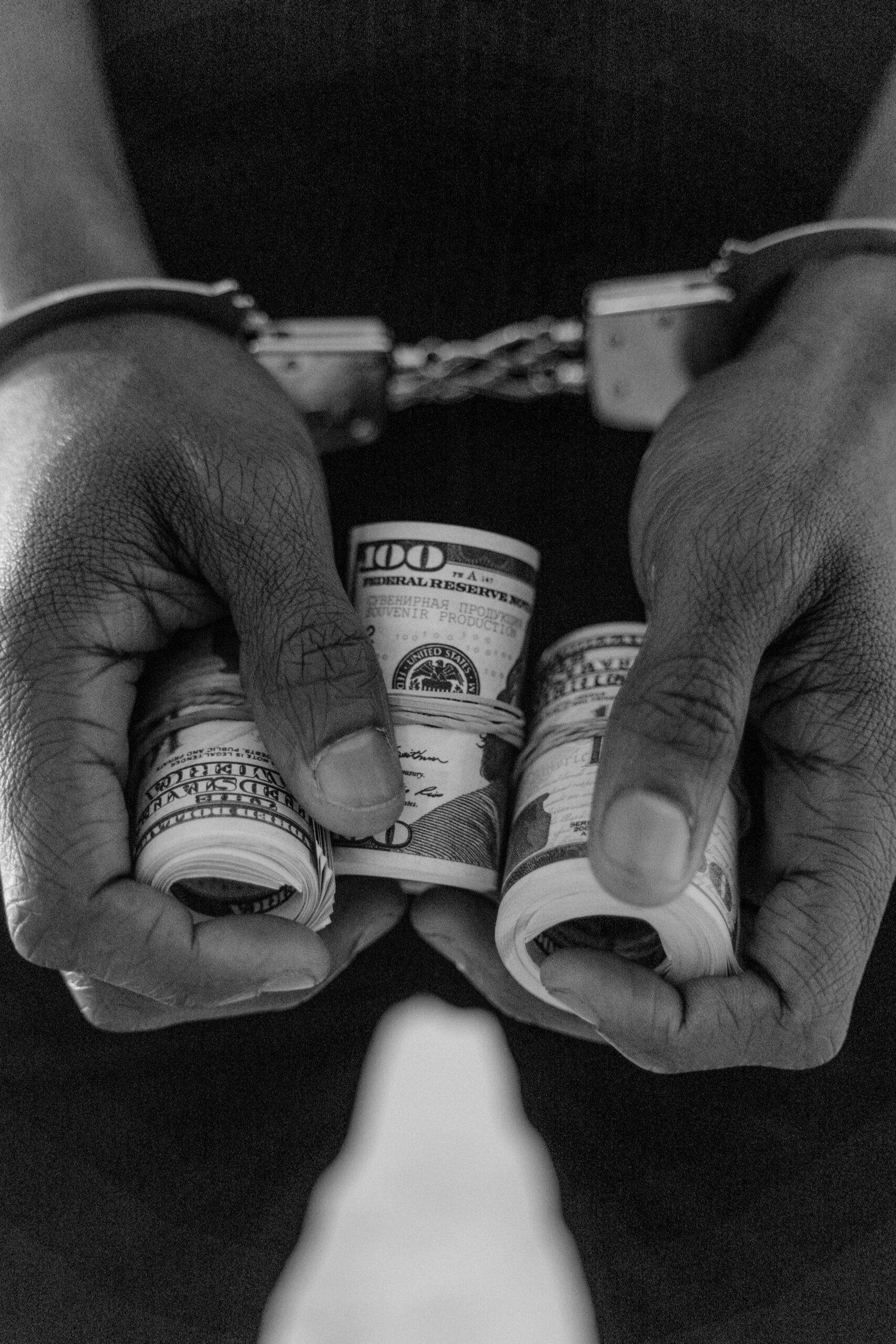Black and white photo of handcuffed hands holding rolls of US dollar bills.